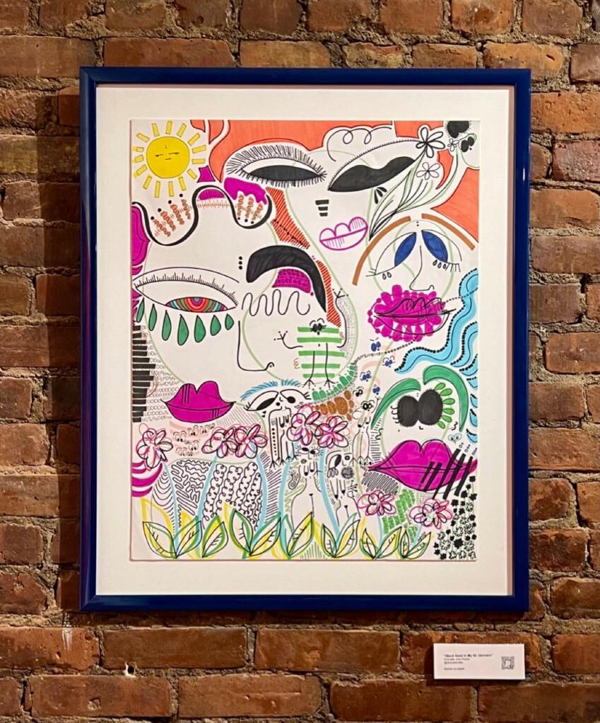 Abstract marker-on-paper artwork Black Sand In My St-Germain by shanelle julia rosita, featuring stylized faces, lips, flowers, and flowing black linework with pink, orange, blue, and green accents, shown framed against a brick wall.